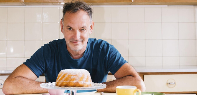 A man is sitting at a table and looking at a juicy Gugelhupf. Cups and saucers are scattered around him on the table. | © Ingo Pertramer