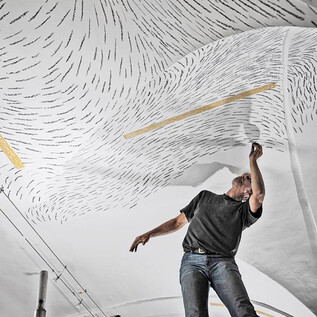 An artist is painting on the ceiling and using black lines on a white background. He is standing on a scaffold and focusing on his work. | © Christoph Müllauer