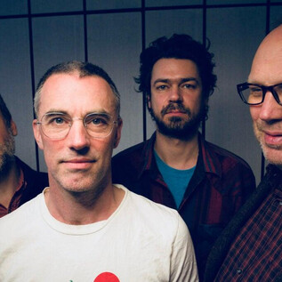 A group of four men stands together in front of a neutral background wall. All have different facial expressions and are wearing different clothes. | © Oliver Potratz