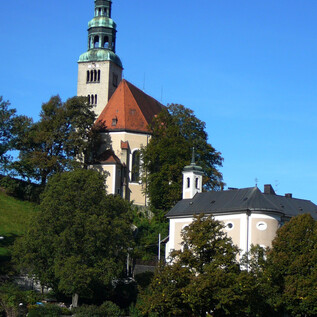Eine Kirche mit einem hohen Turm und einem roten Dach steht auf einem Hügel. Darunter befinden sich einige Bäume und ein kleines Gebäude. | © Pfarre Mülln