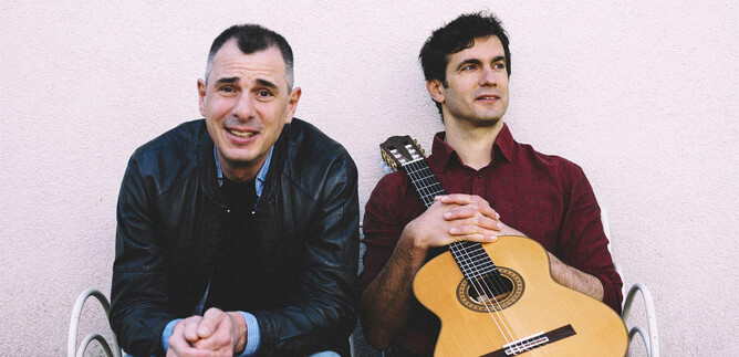 Two men are sitting on a bench, one is smiling, the other has a guitar. They appear relaxed and ready for a musical performance. | © Ingo Pertramer