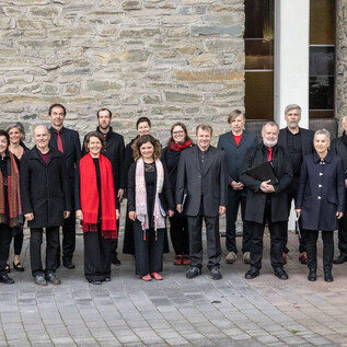 A group of singers stands in front of a stone wall. Most of them are wearing black clothing with red accessories. | © COA
