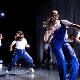 A group of dancers in blue overalls is performing a choreographed presentation on a stage. The mood is energetic and joyful. | © Cornelia Pirchner