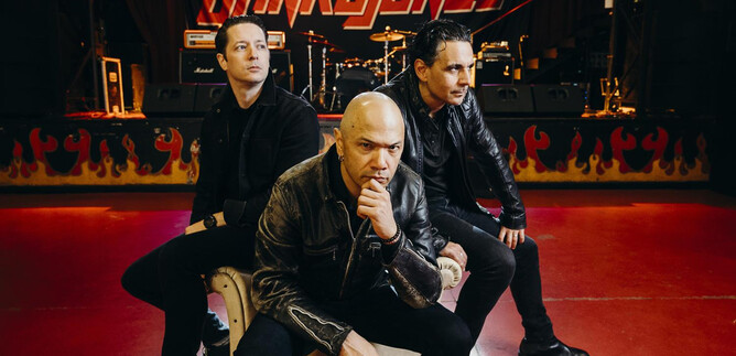 A rock band poses on a chair in a concert hall. In the background, musical instruments and stage lights can be seen. | © Jon_Usual