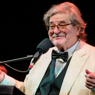 Egy mosolygó idősebb férfi fehér öltönyben és fekete csokornyakkendővel áll a mikrofon előtt. Barátságosnak és szórakoztatónak tűnik a színpadon. | © Karl Satzinger