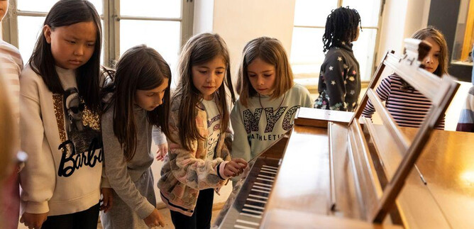 A group of girls is curiously looking at a piano and helping each other. They seem to be having fun exploring the keys. | © Erika Mayer