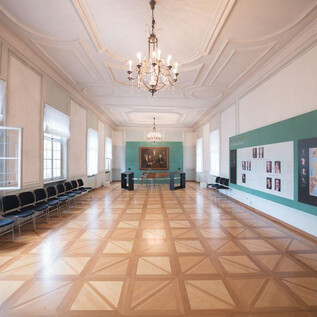 A bright room with an elegant wooden floor and large windows. Pictures and a map are visible on the wall, while chairs are arranged in rows. | © Wolfgang Lienbacher