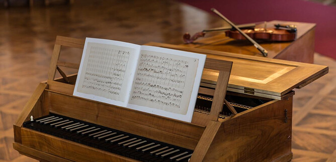 Un piano clásico con una partitura abierta está en primer plano. Al fondo, hay un violín sobre una mesa. | © DomQuartier Salzburg