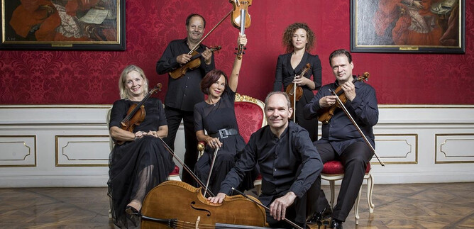 A music group poses in front of a red background with historical paintings. They are playing various string instruments and wearing black clothing. | © DQS/Leopold
