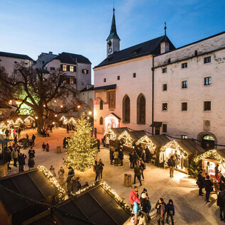 A festive Christmas market with a beautiful tree in the middle. Many people enjoy the atmosphere and the illuminated stalls. | © Festung Hohensalzburg