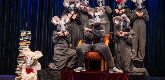 A group of costumed mice stands on a stage, surrounded by a stack of books. In the center, a larger mouse sits on a red throne. | © Leo Fellinger