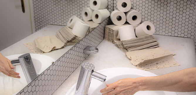 Une salle de bains avec un lavabo, un robinet et des sèche-mains. En arrière-plan, plusieurs rouleaux de papier toilette sont visibles. | © Valentina Schuster