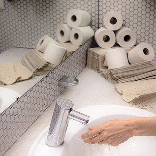 Une salle de bains avec un lavabo, un robinet et des sèche-mains. En arrière-plan, plusieurs rouleaux de papier toilette sont visibles. | © Valentina Schuster