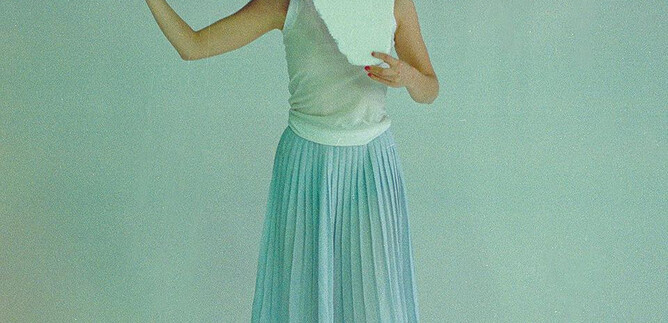 A person is holding a large, rectangular object in front of their face. They are wearing a simple, light-colored skirt and are barefoot on a gentle background. | © Valentina Schuster