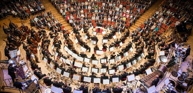 An orchestra plays in a round hall in front of a large audience. The musicians are arranged harmoniously, surrounded by music stands and instruments. | © Holger Talinski