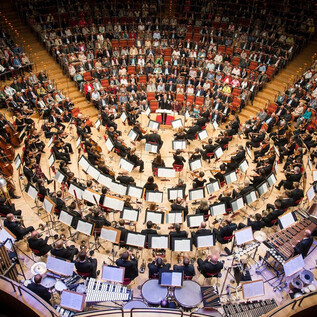 Ein Orchester spielt in einem runden Saal vor einem großen Publikum. Die Musiker sind harmonisch angeordnet, umgeben von Notenständen und Instrumenten. | © Holger Talinski
