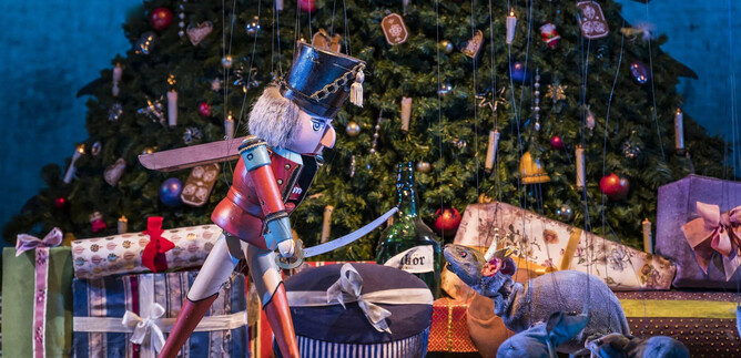 A nutcracker stands in front of a Christmas tree and assisted gifts. Festive decorations can be seen in the background. | © georgeye