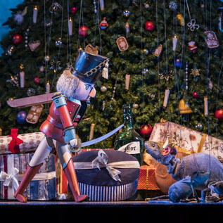 A nutcracker stands in front of a Christmas tree and assisted gifts. Festive decorations can be seen in the background. | © georgeye