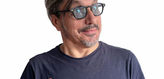 Ein Mann mit grauen Haaren und Brille lächelt und zeigt mit den Händen ein Zeichen der Hoffnung. Er trägt ein einfaches schwarzes T-Shirt und hat einen freundlichen Ausdruck. | © Kian Noam