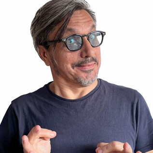 Un homme aux cheveux gris et portant des lunettes sourit et montre un signe d'espoir avec ses mains. Il porte un simple t-shirt noir et a une expression amicale. | © Kian Noam
