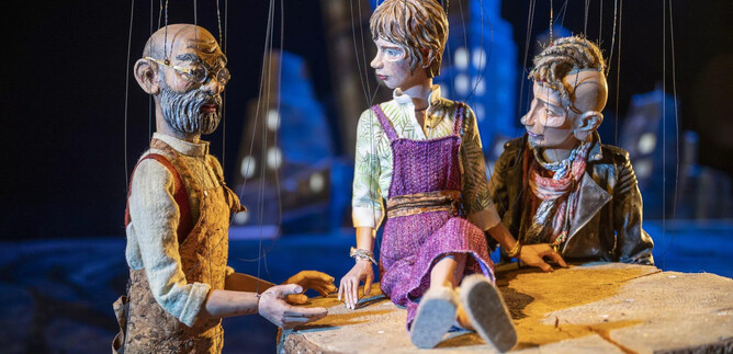 Three dolls are sitting at the edge of a tree stump. The scene feels vibrant with a mystical backlighting. | © Marionettentheater_Bernhard Mueller