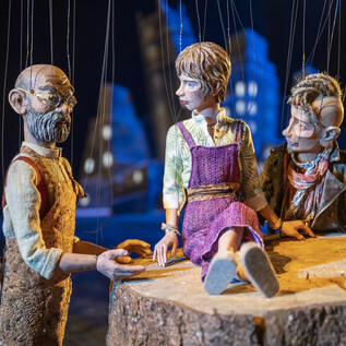 Három baba ül egy fatönk szélén. A jelenet élénknek tűnik egy misztikus háttérvilágítással. | © Marionettentheater_Bernhard Mueller