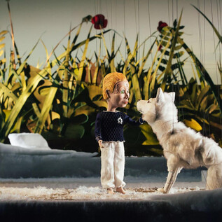 Eine Marionette steht neben einem weißen Wolf in einer lebhaften, grünen Umgebung. Im Hintergrund sind hohe Gräser und rote Blumen zu sehen. | © marco riebler