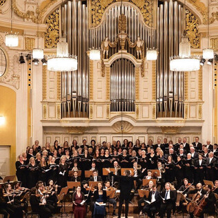Ein großes Orchester und ein gemischter Chor treten auf einer Bühne in einem prächtigen Saal auf. Die Musiker und Sänger sind in festlicher Kleidung und die Atmosphäre ist feierlich. | © Erika Mayer