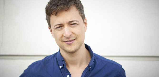 A young man stands in front of a bright wall. He is wearing a blue shirt and smiling kindly at the camera. | © Markus Wache