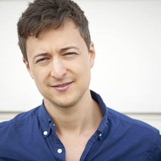 Ein junger Mann steht vor einer hellen Wand. Er trägt ein blaues Hemd und lächelt freundlich in die Kamera. | © Markus Wache