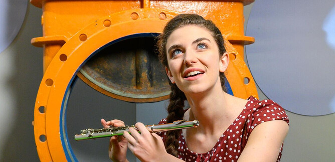 A smiling woman plays the flute in front of an orange background. She is wearing a polka dot dress and looks optimistically to the side. | © SLT / Tobias Witzgall