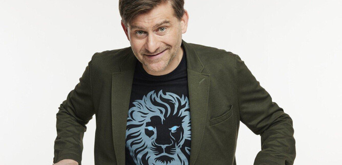 Un hombre con una sonrisa amable lleva una camiseta con un motivo de león. Está sentado a una mesa y tiene un pequeño objeto blanco frente a él. | © Martina Bogdahn