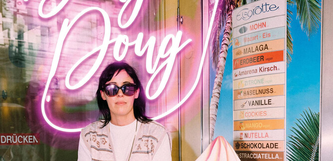 A person is sitting in front of a glowing neon sign with the text "Belly Doug". Next to them is a menu with various ice cream flavors. | © Ingo Pertramer