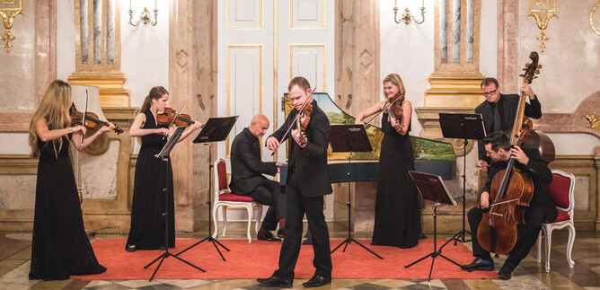 Ein elegantes Musikensemble spielt in einem festlichen Raum. Die Musiker tragen formelle Kleidung und stehen vor Notenständern. | © Michael Groessinger