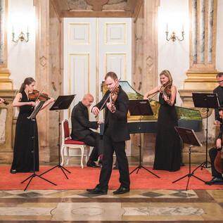 Ein elegantes Musikensemble spielt in einem festlichen Raum. Die Musiker tragen formelle Kleidung und stehen vor Notenständern. | © Michael Groessinger
