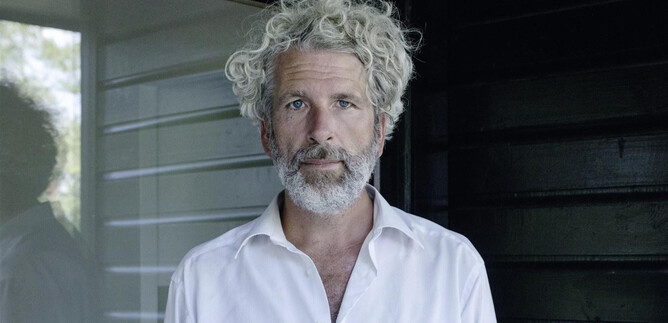 A man with curly, gray hair stands in front of a dark wall. He is wearing a white shirt and has a thoughtful expression. | © Gerald von Foris