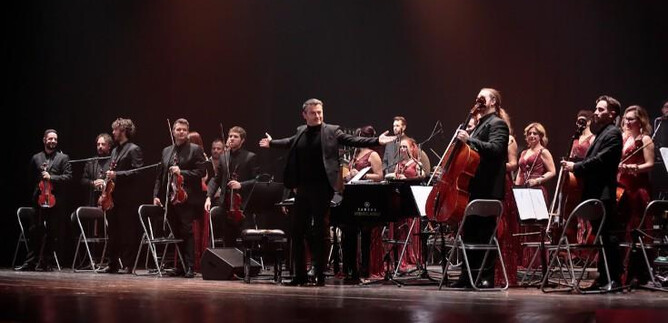 Ein Orchester steht auf der Bühne und spielt. Der Dirigent leitet die Musiker, die in eleganten Kleidern gekleidet sind. | © The Showtime Agency GmbH