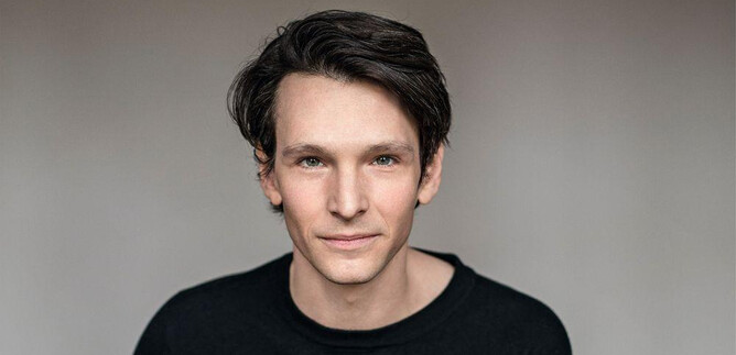 Egy fiatalember sötét, göndör hajjal fekete felsőt visel. Barátságosan néz a kamerába egy semleges háttér előtt. | © Sabine Höhn