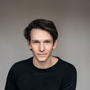 A young man with dark, curly hair is wearing a black top. He is looking kindly into the camera against a neutral background. | © Sabine Höhn