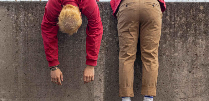 Two people are hanging upside down over a concrete wall. They are wearing red and beige clothing and appear relaxed. | © Schauspielhaus Salzburg