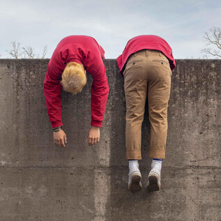 Dos personas están colgadas cabeza abajo sobre un muro de concreto. Llevan ropa roja y beige y parecen relajadas. | © Schauspielhaus Salzburg