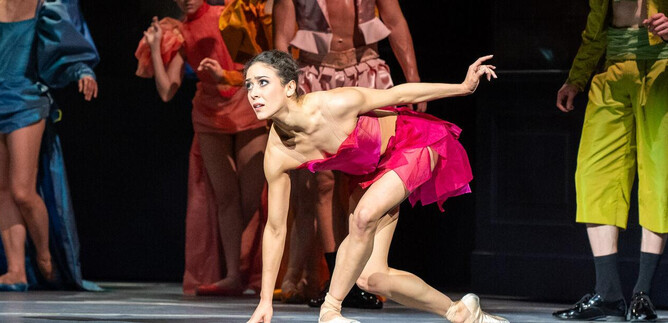 A dancer in a pink outfit moves dynamically on stage. In the background, other dancers in colorful costumes can be seen. | © SLT / Tobias Witzgall