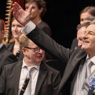 Ein Musiker in einem schwarzen Anzug lächelt und winkt zum Publikum. Im Hintergrund sind weitere Orchestermitglieder mit Instrumenten zu sehen. | © Christian Schneider