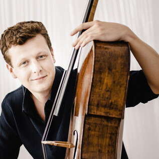 A young man poses with a cello. He has a friendly smile and is wearing a black shirt. | © Marco_Borggreve