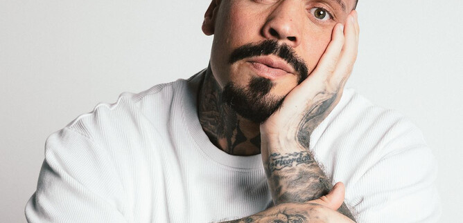 A man with many tattoos looks thoughtfully at the camera. He is wearing a white T-shirt and has a beard. | © Ramiro Simone