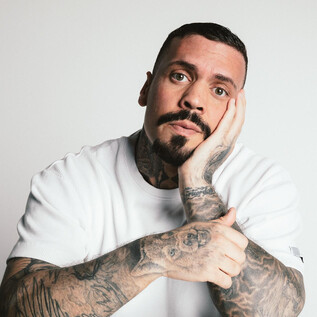 Mężczyzna z wieloma tatuażami patrzy zagubionym wzrokiem w kamerę. Ma na sobie białą koszulkę i brodę. | © Ramiro Simone