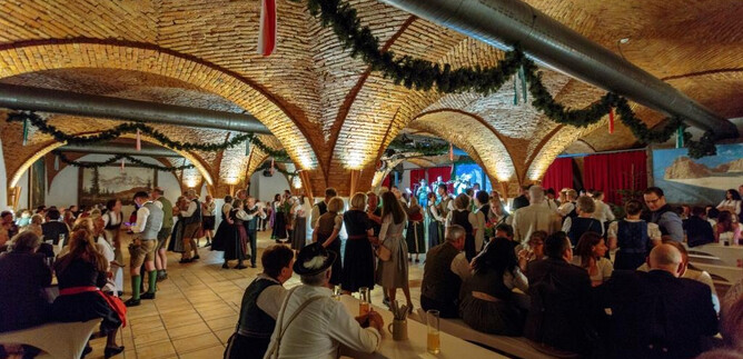 Eine lebhafte Veranstaltung in einem festlich dekorierten Raum mit gewölbten Decken. Menschen in traditioneller Kleidung tanzen und feiern zusammen. | © Edelweiss-Club