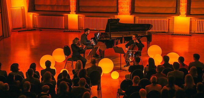 Zespół muzyczny z fortepianem, wiolonczelą i skrzypcami gra przed publicznością w ciepło oświetlonym pomieszczeniu. Wokół muzyków umieszczone są duże, świecące kule. | © @Freie Kunst