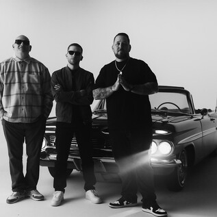 Drei Männer stehen vor einem klassischen Auto in einem minimalistischen Studiobereich. Die Szene ist schwarz-weiß und vermittelt einen starken visuellen Eindruck. | © Rockhouse Salzburg