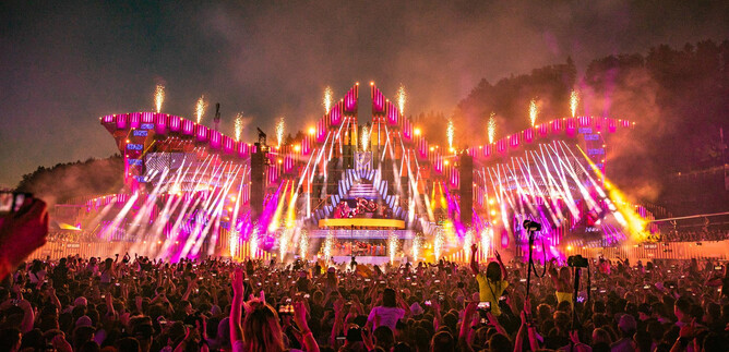 Ein lebhaftes Musikfestival mit einer riesigen Bühne, die von bunten Lichtern und Feuerwerk umgeben ist. Die Menge jubelt und ist voller Energie. | © Geoffrey Hubbel
